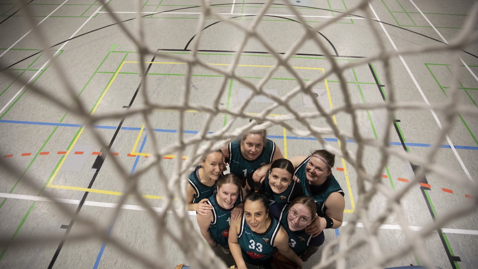 Frauen in einer Sporthalle, die durch einen Basketballkorb nach oben den Betrachter ansehen.
