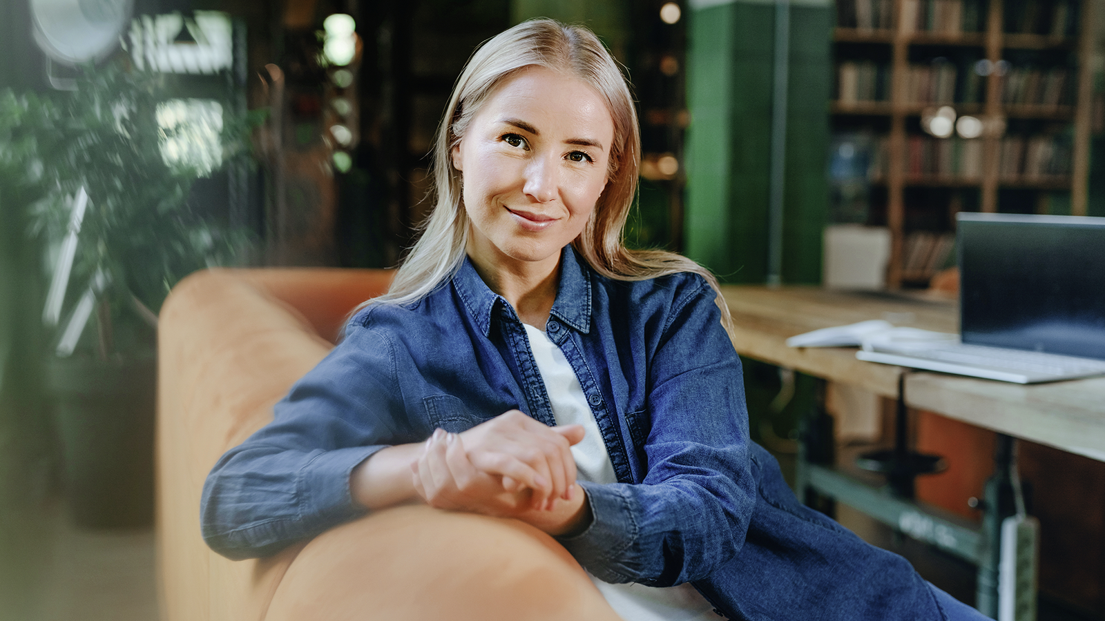 Blonde Frau in Jeanshemd sitzt entspannt auf einem Sofa in einem modernen Büro- oder Arbeitsbereich und blickt freundlich in die Kamera.