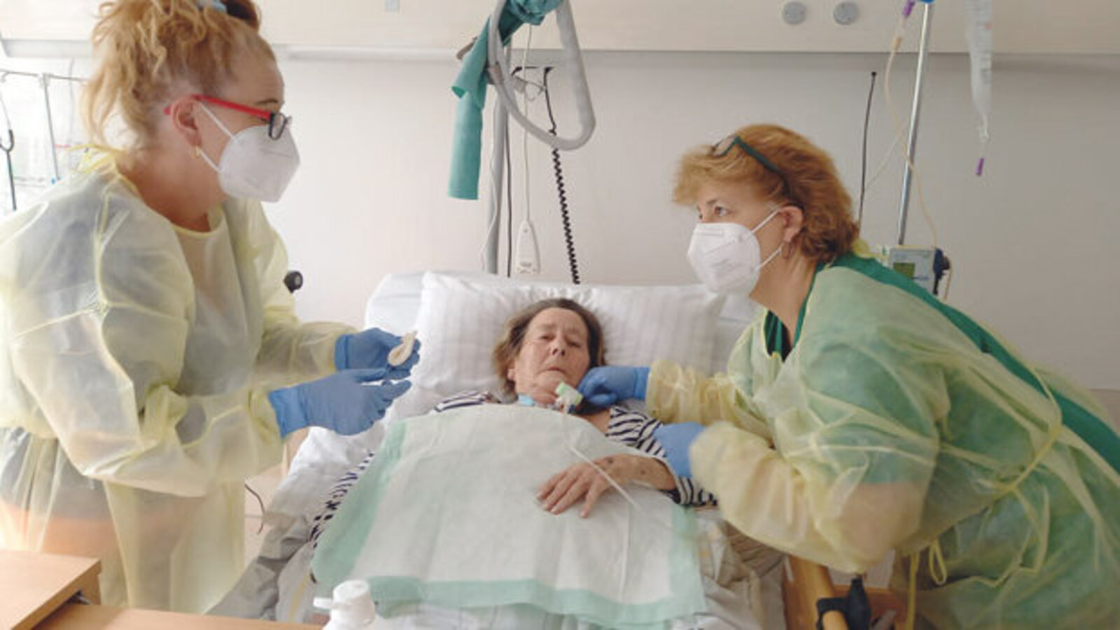 Drei Frauen, zwei in Schutzkleidung an einem Krankenhausbett, dritte Frau im Bett.