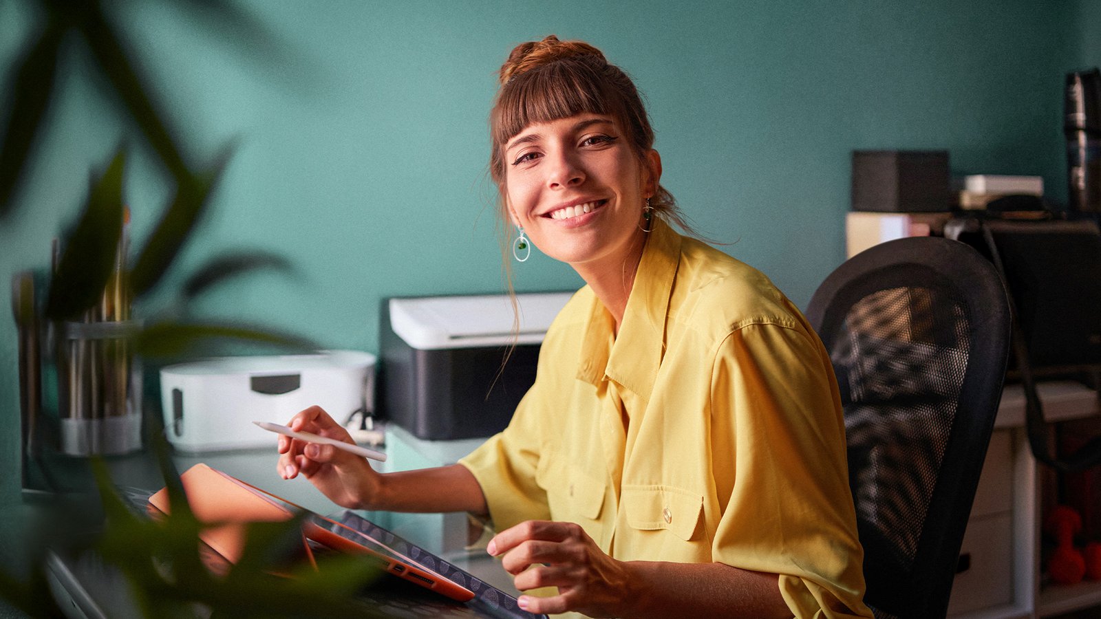 Frau sitzt am Schreibtisch, lächelt in die Kamera und arbeitet mit Tablet und Eingabestift in einem modernen Arbeitsumfeld.