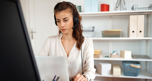 Callcenter Mitarbeiterin mit headset schaut lächelnd in die Kamera, im Hintergrund sind zwei weitere Mitarbeiterinnen.