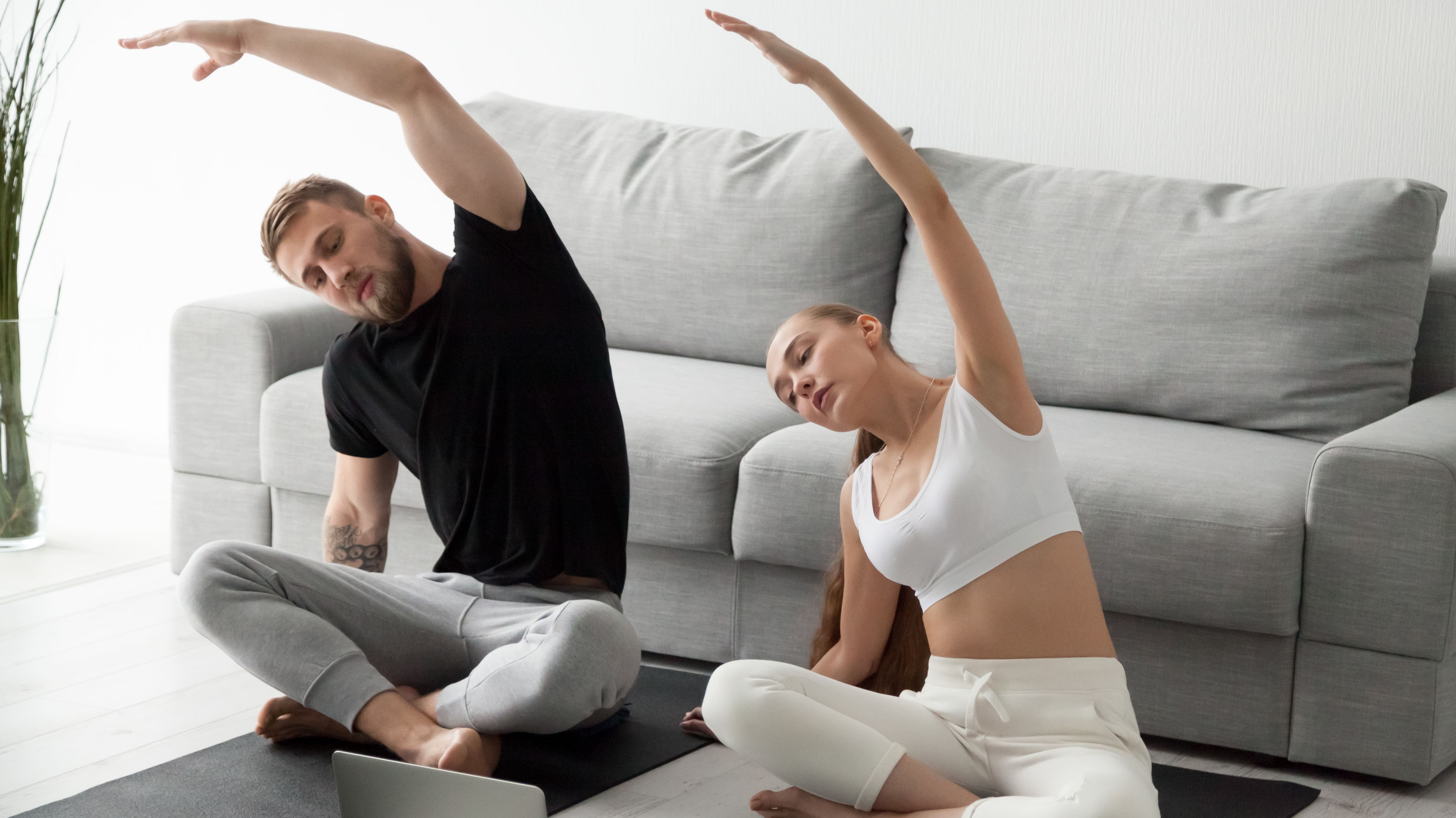 Mann und Frau machen zu Hause Yoga-Übungen und schauen dabei auf ein Laptop.