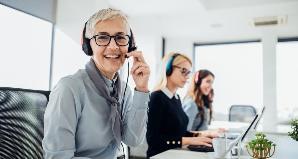 Callcenter Mitarbeiterin mit headset schaut lächelnd in die Kamera, im Hintergrund sind zwei weitere Mitarbeiterinnen.