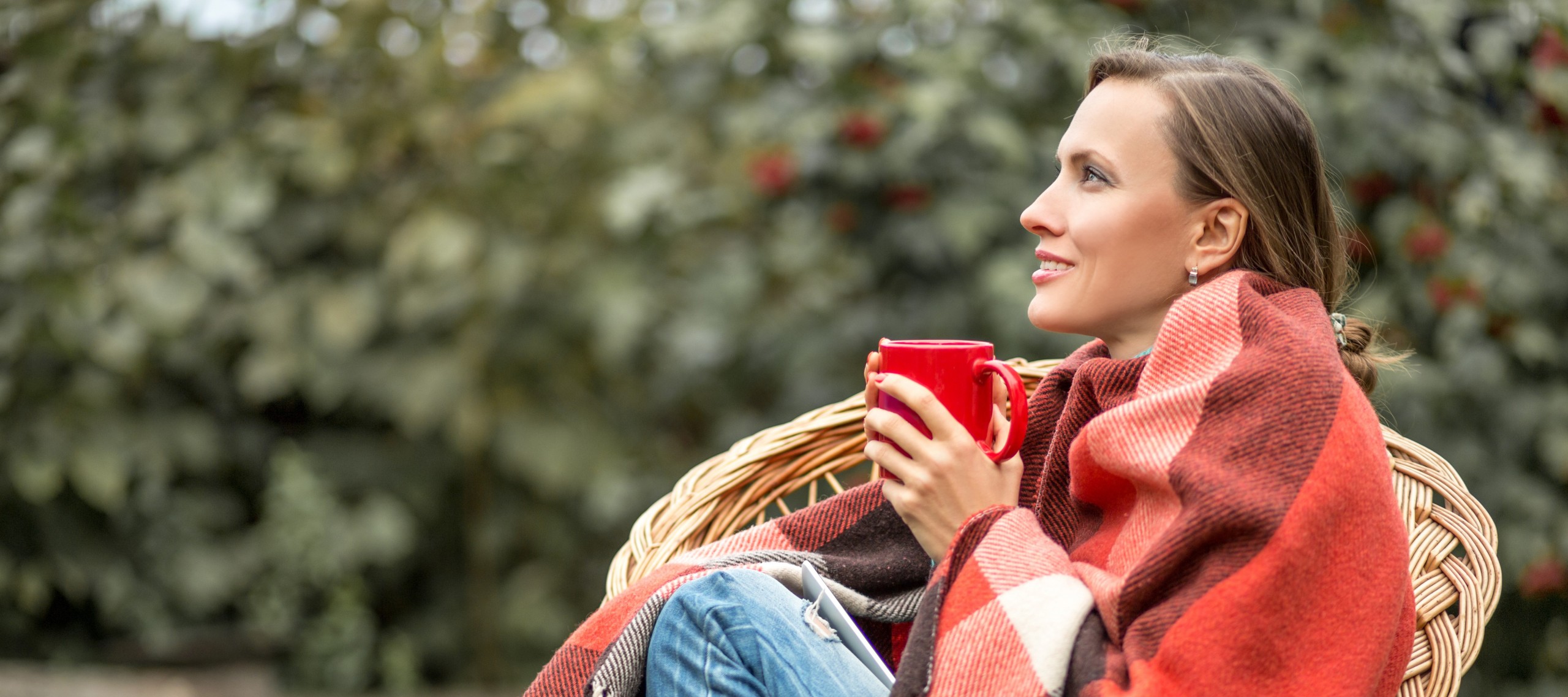 Frau sitzt draußen auf einem Rattanstuhl, eingehüllt in eine Decke, mit einer roten Tasse in beiden Händen.