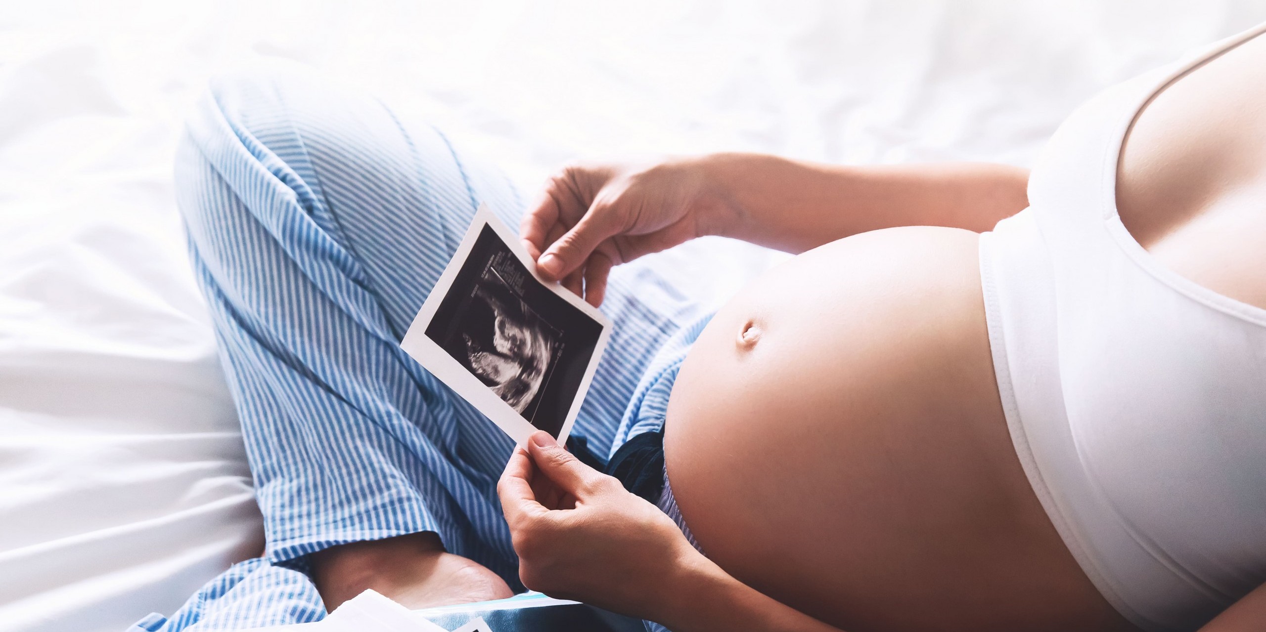 Hochschwangere Frau sitzt im Schneidersitz auf dem Bett und betrachtet Ultraschallbilder vom Kind.