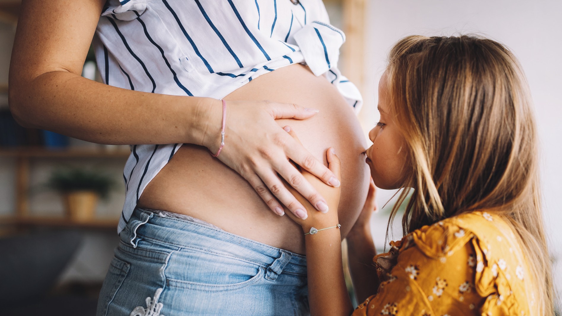 Hochschwangere Frau steht mit Babybauch vor ihrer kleinen Tochter und diese gibt einen Kuss auf den Bauch. 