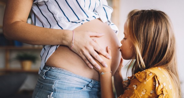 Hochschwangere Frau steht mit Babybauch vor ihrer kleinen Tochter und diese gibt einen Kuss auf den Bauch. 