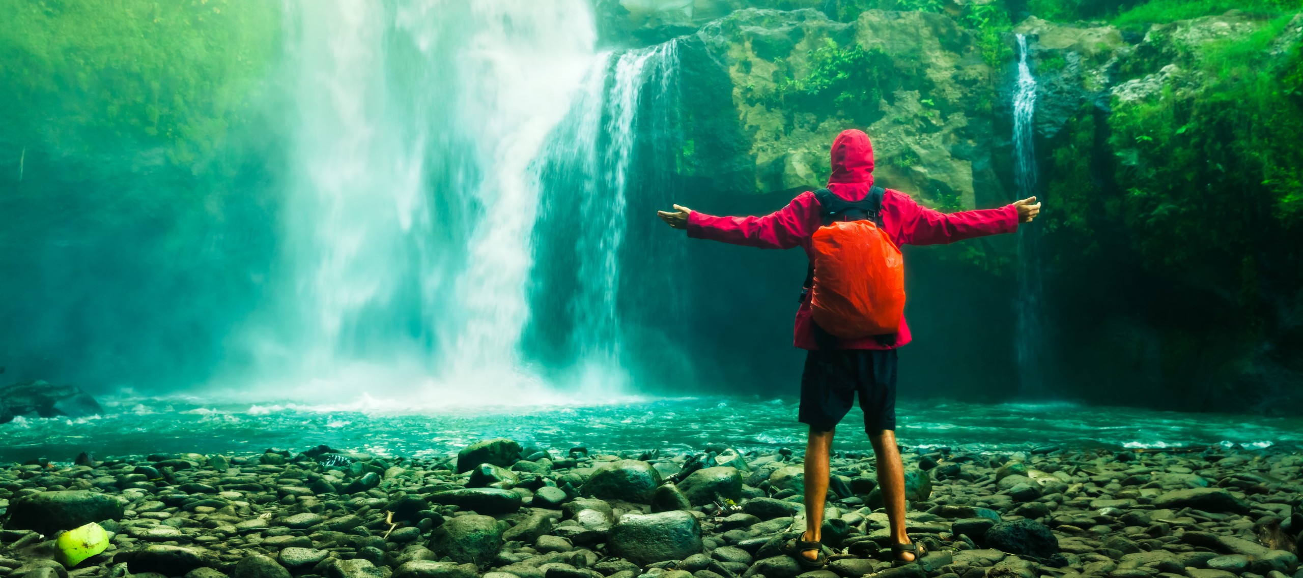 	Mann steht mit ausgebreiteten Armen im Regencape und kurzer Hose mit dem Rücken zur Kamera vor einem tropischen Wasserfall.