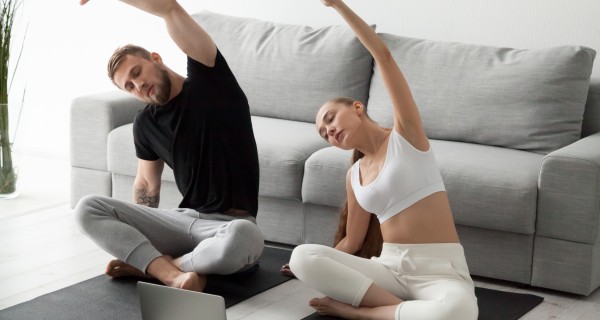 Mann und Frau machen zu Hause Yoga-Übungen und schauen dabei auf ein Laptop.