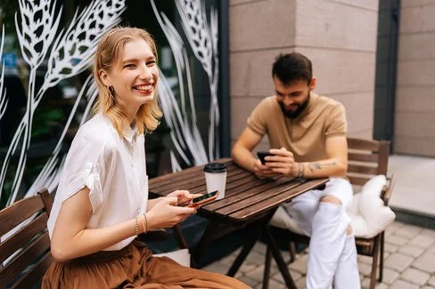 Ein junges Paar sitzt draußen an einem Tisch eines Cafés. Beide halten ihr Handy in der Hand und lächeln.