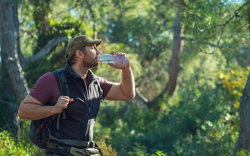 Ein Mann trinkt beim Wandern im Wald Wasser aus einer Flasche, um Kopfschmerzen vorzubeugen.
