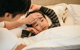 Ein Vater fasst seinem Sohn an die Stirn, der mit Pfeifferschem Drüsenfieber im Bett liegt.