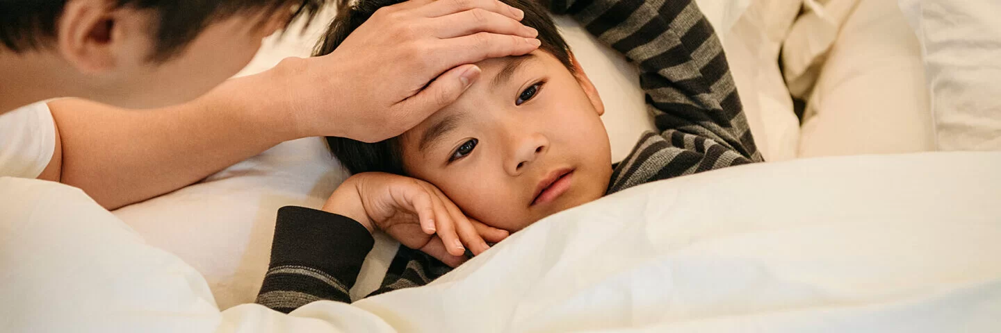 Ein Vater fasst seinem Sohn an die Stirn, der mit Pfeifferschem Drüsenfieber im Bett liegt.