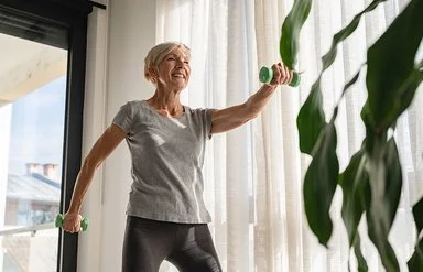 Eine ältere Frau steht in einem Wohnzimmer und treibt Sport. In beiden Händen hält sich eine Hantel, mit denen sie ihre Muskulatur kräftigt.