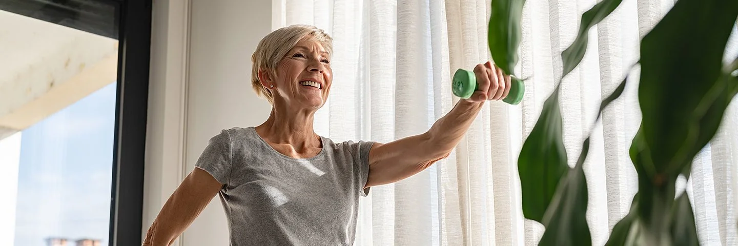 Eine ältere Frau steht in einem Wohnzimmer und treibt Sport. In beiden Händen hält sich eine Hantel, mit denen sie ihre Muskulatur kräftigt.
