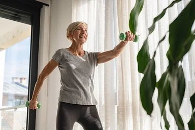 Eine ältere Frau steht in einem Wohnzimmer und treibt Sport. In beiden Händen hält sich eine Hantel, mit denen sie ihre Muskulatur kräftigt.