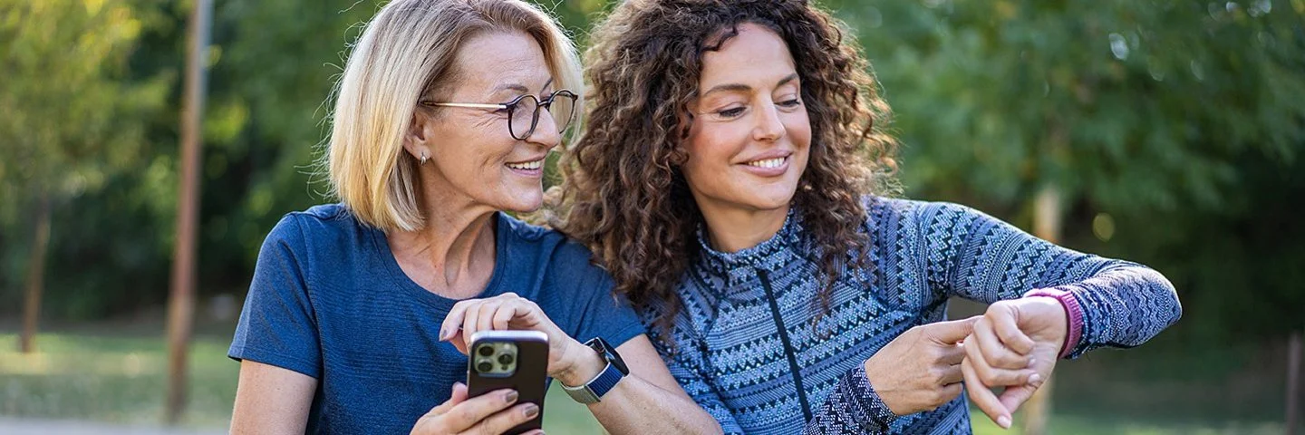 Zwei Frauen sitzen in Sportkleidung auf Sportmatten in einem grünen Park und betrachten Ergebnisse auf ihren Smartwatches.
