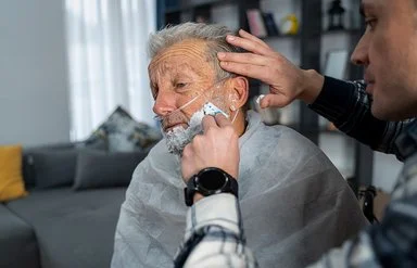 Einem pflegebedürftigen Mann mit einem Beatmungsgerät bekommt zu Hause von einer männlichen Person das Gesicht rasiert.