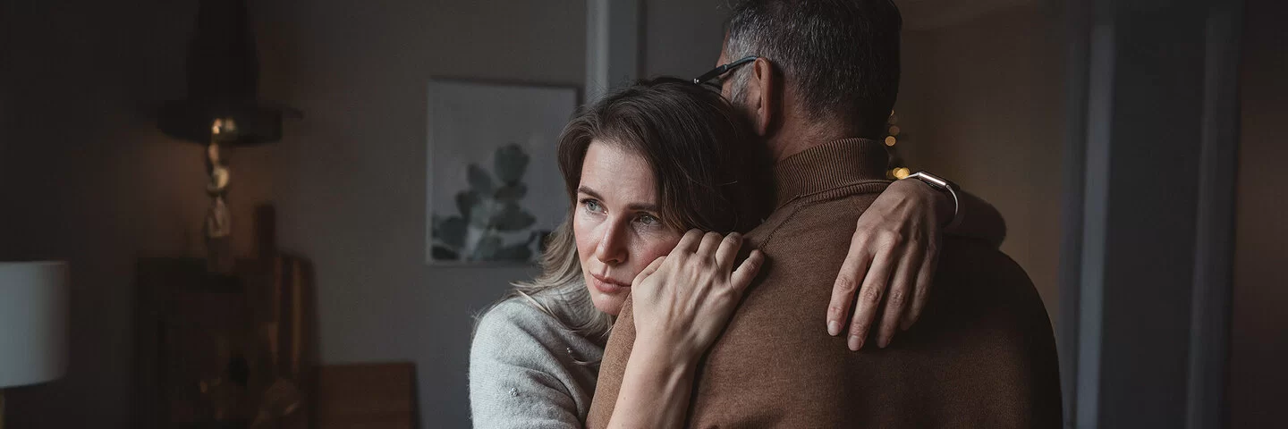 Ein Paar in den mittleren Jahren umarmt sich. Der Mann ist nur von hinten zu sehen, die Frau schaut traurig und nachdenklich zur Seite.