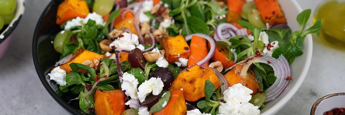 Kürbissalat mit Feta nach einem Rezept von Felicitas Then, angerichtet auf einem schwarz-weißen Teller, garniert mit Weintrauben und Zwiebeln.