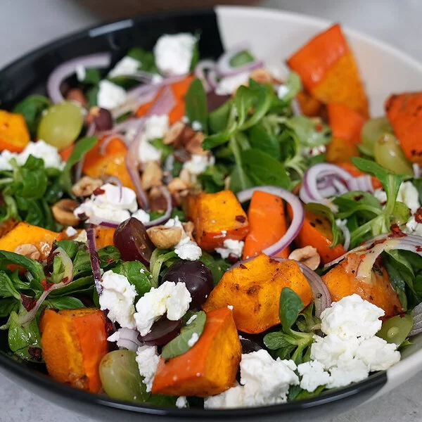 Kürbissalat mit Feta nach einem Rezept von Felicitas Then, angerichtet auf einem schwarz-weißen Teller, garniert mit Weintrauben und Zwiebeln.