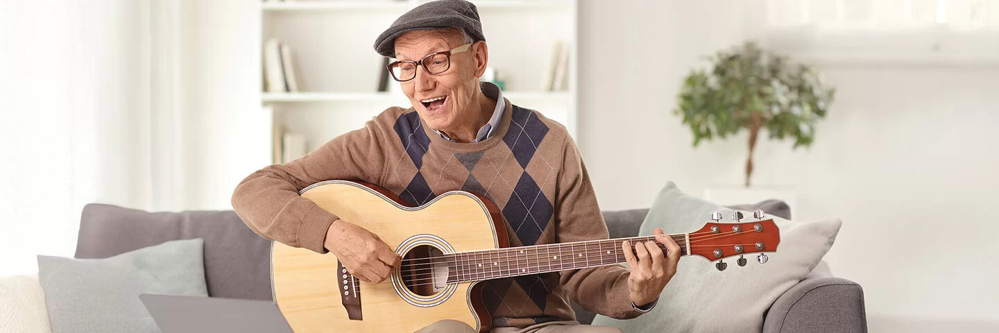 Lebenslanges Lernen: Ein älterer Mann sitzt auf dem Sofa vor dem Laptop und übt auf einer akustischen Gitarre.
