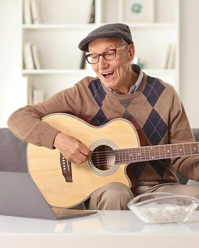 Lebenslanges Lernen: Ein älterer Mann sitzt auf dem Sofa vor dem Laptop und übt auf einer akustischen Gitarre.