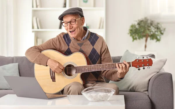 Lebenslanges Lernen: Ein älterer Mann sitzt auf dem Sofa vor dem Laptop und übt auf einer akustischen Gitarre.