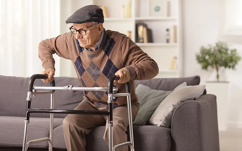Älterer Mann mit Schiebermütze und Pullover auf einem Sofa. Vor ihm steht eine Gehhilfe. Er stützt sich auf den Rollator um aufzustehen.