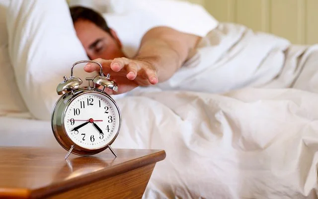 Ein Mann im Bett greift nach einem Wecker, der auf dem Nachttisch steht.