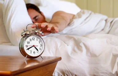 Ein Mann im Bett greift nach einem Wecker, der auf dem Nachttisch steht.