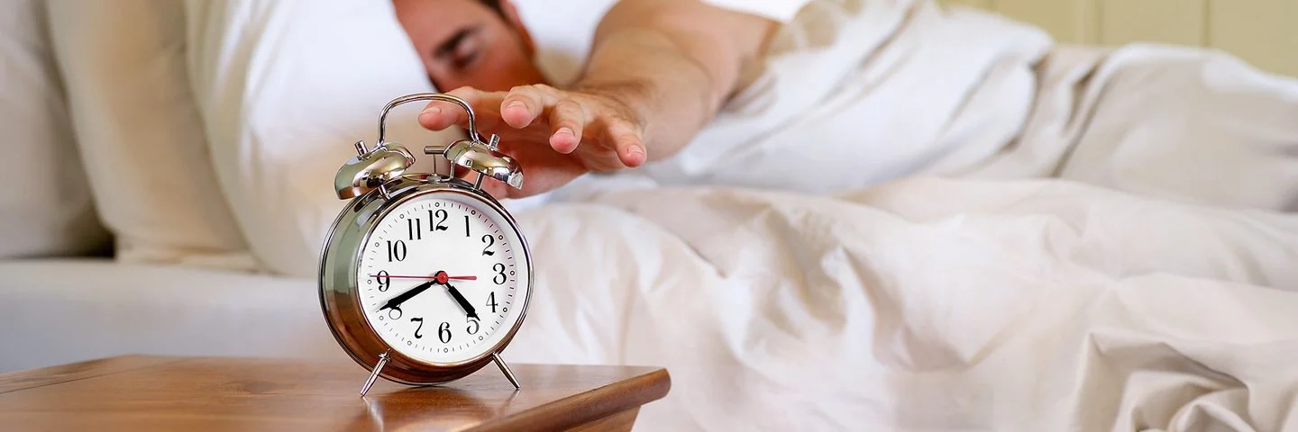 Ein Mann im Bett greift nach einem Wecker, der auf dem Nachttisch steht.