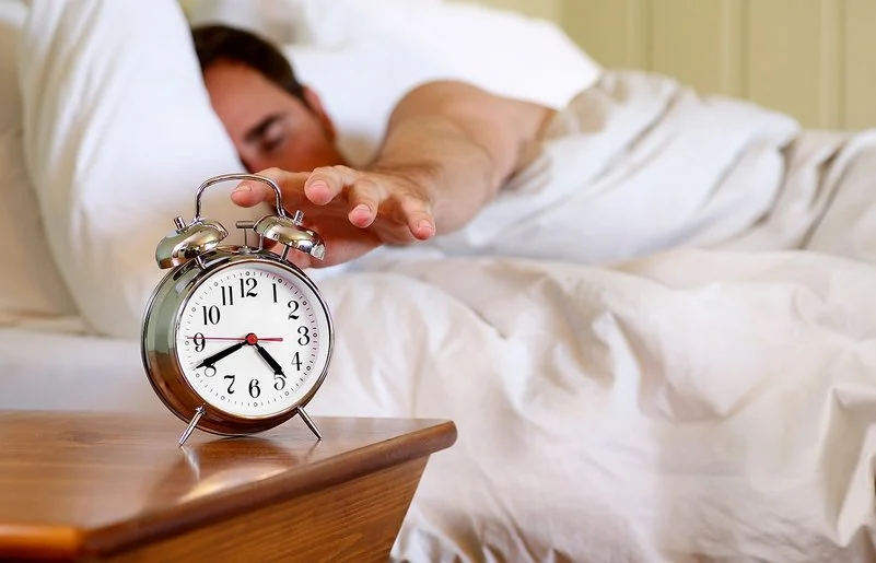 Ein Mann im Bett greift nach einem Wecker, der auf dem Nachttisch steht.