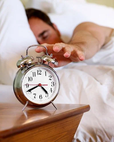 Ein Mann im Bett greift nach einem Wecker, der auf dem Nachttisch steht.