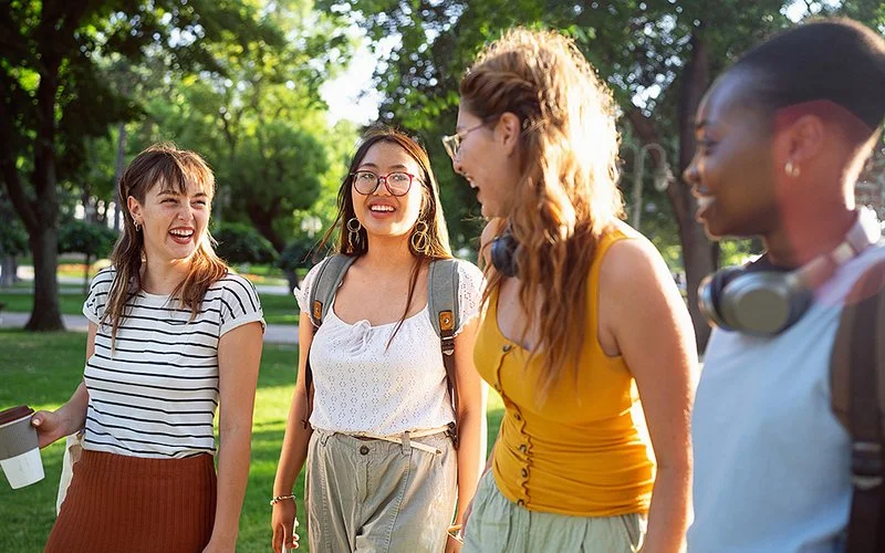 Vier Frauen gehen lachend nebeneinander her und unterhalten sich. Die Frauen unterscheiden sich in Statur, Haarfarbe, Hautfarbe und Kleidungsstil und scheinen freundschaftlich miteinander verbunden zu sein.