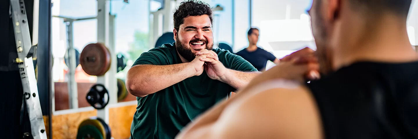 Ein lachender Mann mit Übergewicht powert sich im Fitnessstudio aus. Gemeinsam mit einem Personaltrainer trainiert er Squats. Im Hintergrund sind Geräte mit Hanteln sowie wie ein weiterer Mann zu sehen.