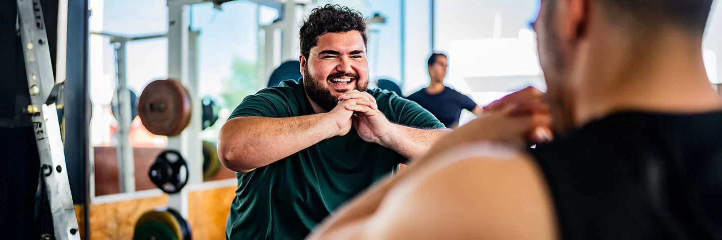 Ein lachender Mann mit Übergewicht powert sich im Fitnessstudio aus. Gemeinsam mit einem Personaltrainer trainiert er Squats. Im Hintergrund sind Geräte mit Hanteln sowie wie ein weiterer Mann zu sehen.