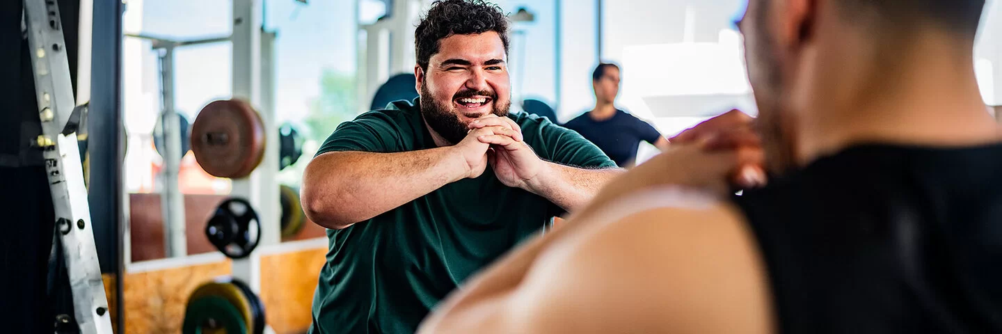 Ein lachender Mann mit Übergewicht powert sich im Fitnessstudio aus. Gemeinsam mit einem Personaltrainer trainiert er Squats. Im Hintergrund sind Geräte mit Hanteln sowie wie ein weiterer Mann zu sehen.