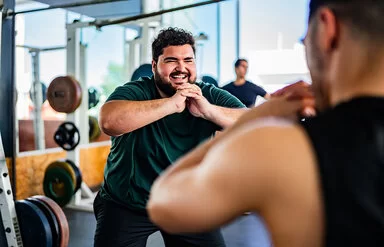 Ein lachender Mann mit Übergewicht powert sich im Fitnessstudio aus. Gemeinsam mit einem Personaltrainer trainiert er Squats. Im Hintergrund sind Geräte mit Hanteln sowie wie ein weiterer Mann zu sehen.