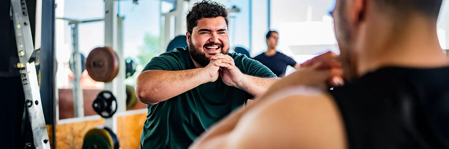 Ein lachender Mann mit Übergewicht powert sich im Fitnessstudio aus. Gemeinsam mit einem Personaltrainer trainiert er Squats. Im Hintergrund sind Geräte mit Hanteln sowie wie ein weiterer Mann zu sehen.