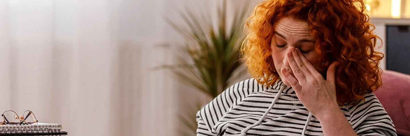 Junge Frau mit gestreiftem Sweatshirt liest auf einer Couch in einer Broschüre und reibt sich die Augen.