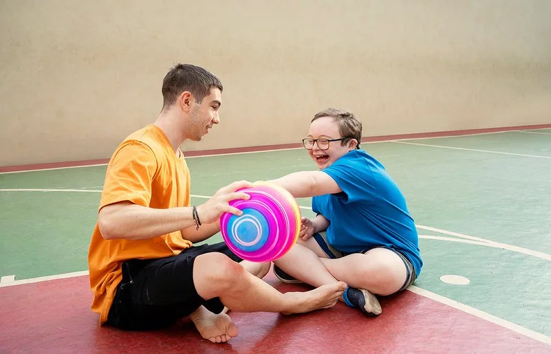 Zwei männliche Jugendliche sitzen auf dem Boden einer Turnhalle und spielen mit einem Ball. Einer von ihnen hat das Down-Syndrom.