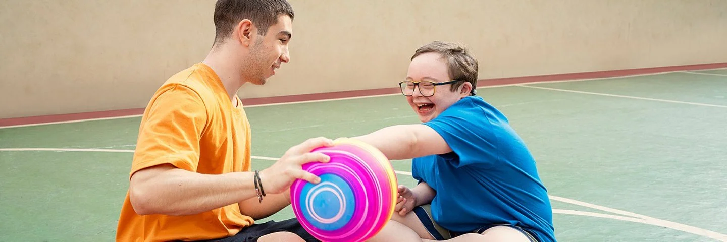 Zwei männliche Jugendliche sitzen auf dem Boden einer Turnhalle und spielen mit einem Ball. Einer von ihnen hat das Down-Syndrom.