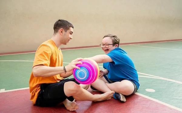 Zwei männliche Jugendliche sitzen auf dem Boden einer Turnhalle und spielen mit einem Ball. Einer von ihnen hat das Down-Syndrom.