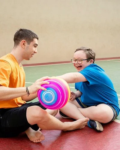 Zwei männliche Jugendliche sitzen auf dem Boden einer Turnhalle und spielen mit einem Ball. Einer von ihnen hat das Down-Syndrom.