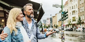 Eine blonde Frau mittleren Alters und ein Mann mit grauen Haaren und Vollbart stehen in einer Stadt. Sie lächelt und schaut dabei nach links während er geradeaus an ihr vorbeischaut.