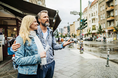 Eine blonde Frau mittleren Alters und ein Mann mit grauen Haaren und Vollbart stehen in einer Stadt. Sie lächelt und schaut dabei nach links während er geradeaus an ihr vorbeischaut.