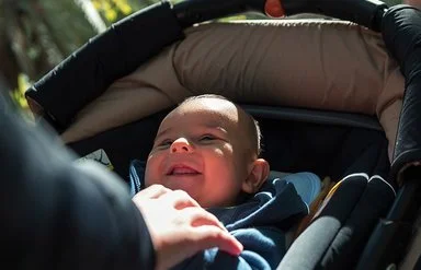 Ein Baby liegt in einem Kinderwagen und schaut nach oben, während der Wagen auf einem sonnigen Waldweg geschoben wird.