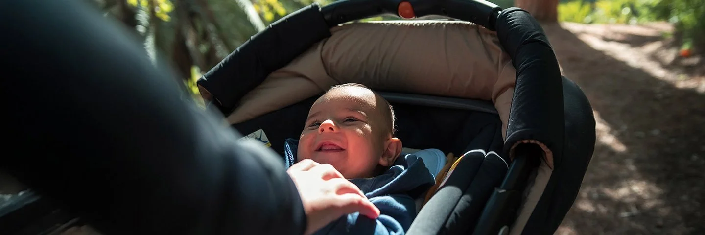 Ein Baby liegt in einem Kinderwagen und schaut nach oben, während der Wagen auf einem sonnigen Waldweg geschoben wird.