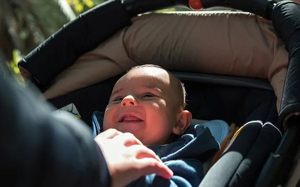 Ein Baby liegt in einem Kinderwagen und schaut nach oben, während der Wagen auf einem sonnigen Waldweg geschoben wird.